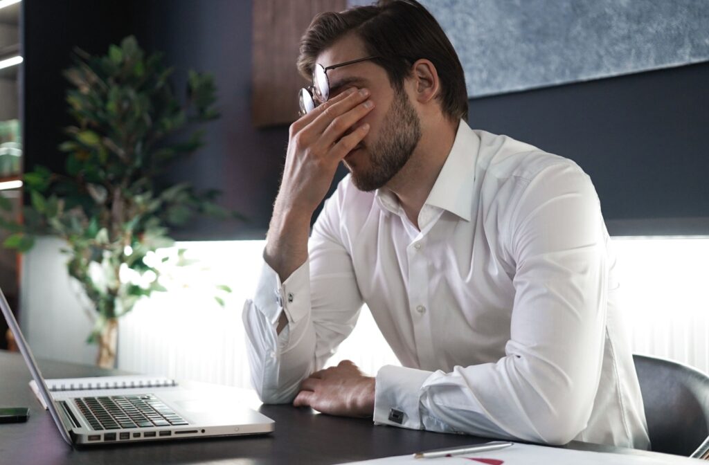 A person in a white shirt sits at a desk with a laptop, rubbing their eyes in discomfort.