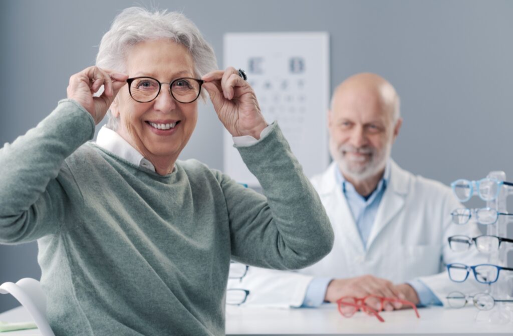 A mature patient cheerfully shows off their new eyeglasses while their optometrist sits in the background.
