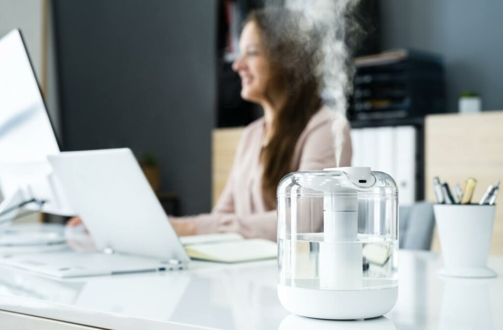 A desktop humidifier releasing mist on an office desk while a person works on a laptop in the background.