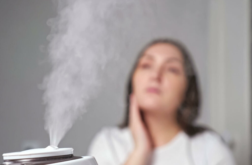 A humidifier releasing visible mist in the foreground with a person sitting in the background, lightly touching their face.